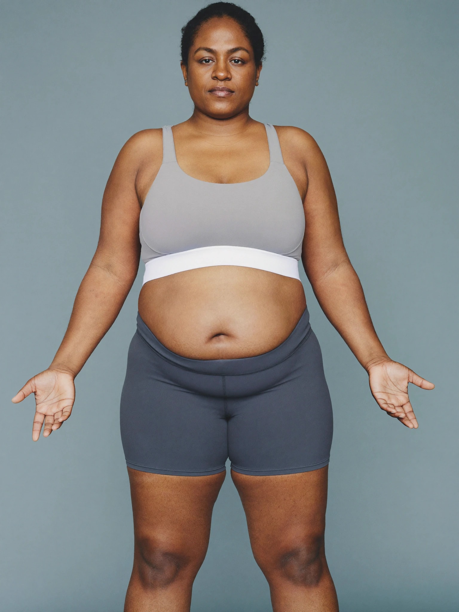 A woman standing facing the camera in a sports bra and shorts, with the stomach, front of the thighs and upper arms highlighted as injection sites
