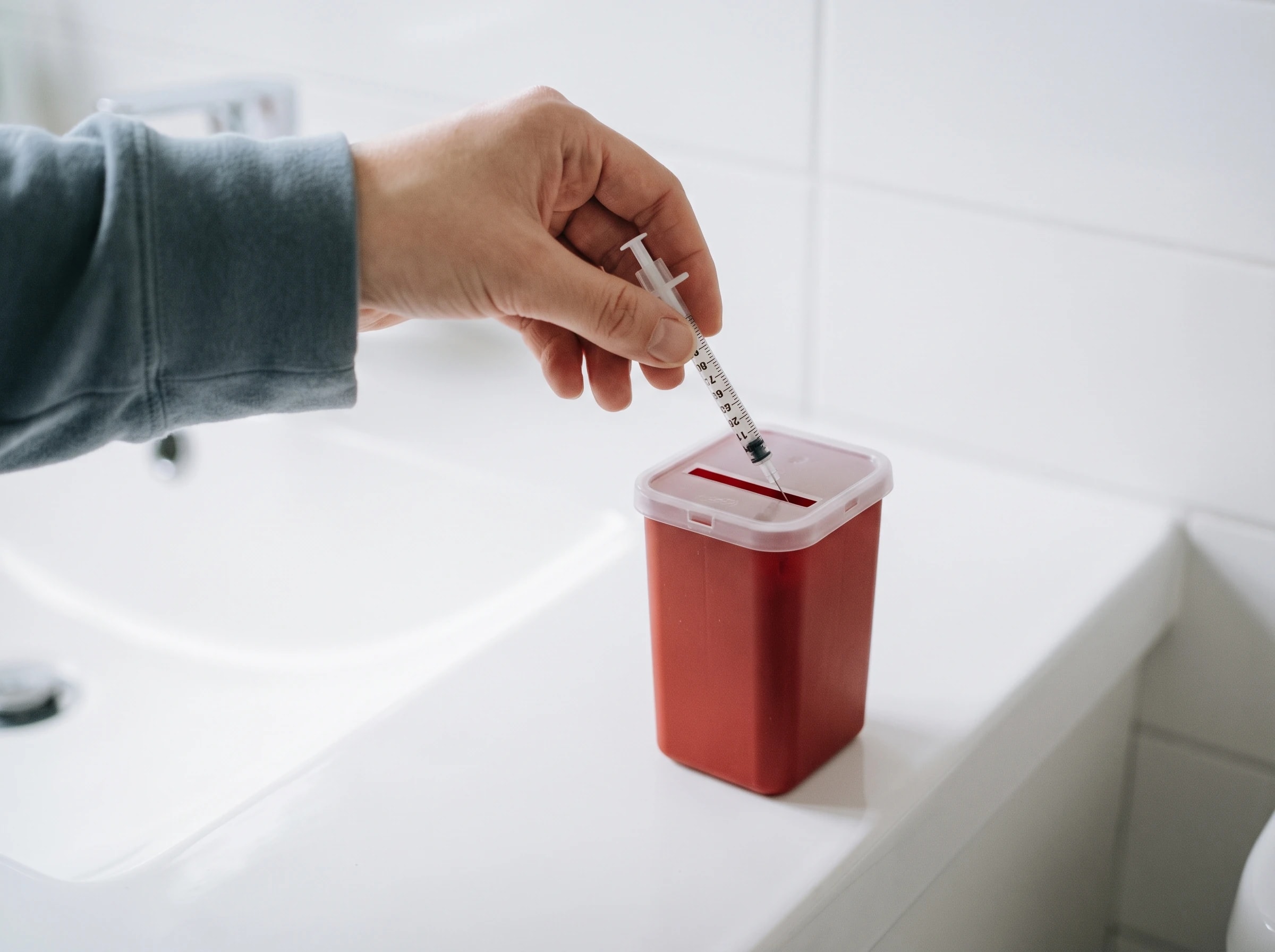 A hand dropping a used insulin syringe into a red sharps container